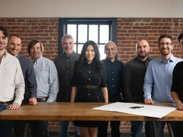 Nine people stand behind a wooden table with paper and pens, in a room with brick walls and a window.